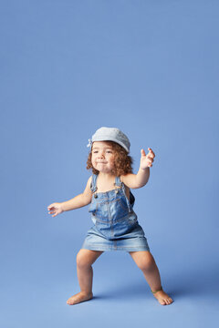 Naklejki Charming barefoot child in denim dress and hat with curly hair looking away while dancing on blue background