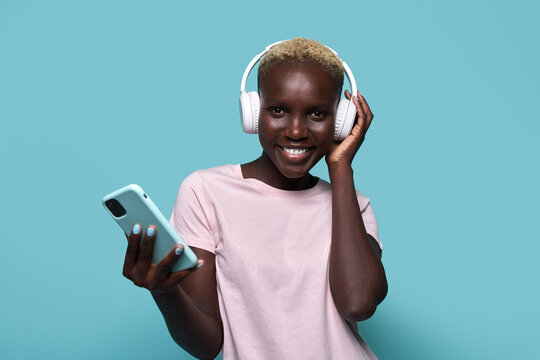 Cheerful African American female toothy smiling looking at camera while listening to music in headphones against blue background