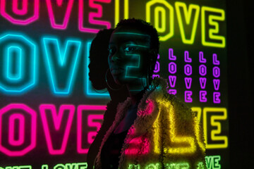 African American woman in trendy fur coat looking at camera while standing near dark wall with bright neon Love inscriptions