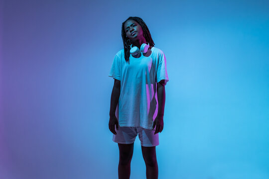 Serious African American teenage girl in headphones on neck standing in studio with bright glowing neon lights