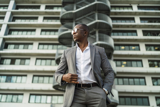 From below of adult well dressed black male executive in sunglasses looking away against multistory house in city