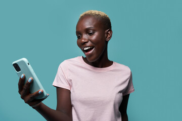 Expressive beautiful African American female with short hair and bright manicure browsing on smartphone against blue background