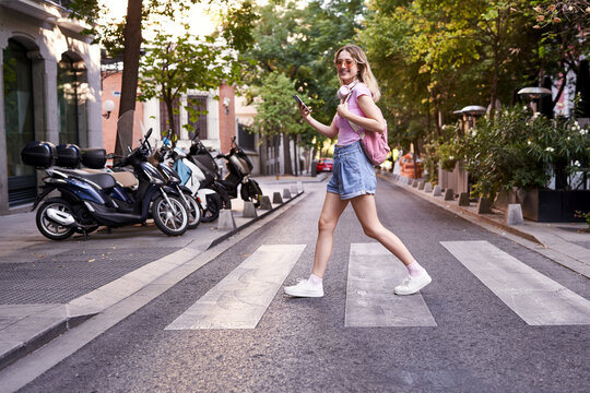 Side view full body of young woman walking on pedestrian crossing and checking route on mobile phone in Madrid