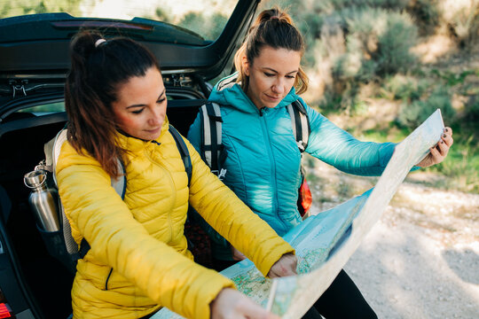 Content female hikers sitting on car trunk and navigating on paper map while searching for route and pointing away