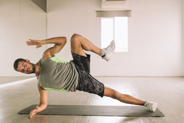 Full body of young focused fit determined sportsman in activewear performing High Side Plank with leg raises exercise during workout in studio near big wall mirror
