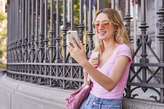Side view of positive teenage female with backpack smiling brightly while scrolling mobile phone leaning on metal fence