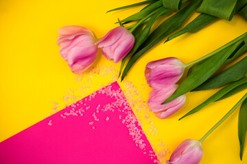 Pink tulips with green leaves in the upper right corner on a yellow background. Pink box for text. Salt crystals are scattered over the background. View from above. Postcard, poster. 