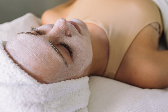 High angle of young calm female client with towel on head lying on couch with closed eyes and white facial mask and relaxing during skincare procedure