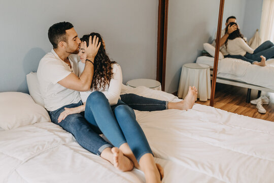 Loving couple sitting on soft bed in bedroom and tenderly hugging while enjoying weekend together at home