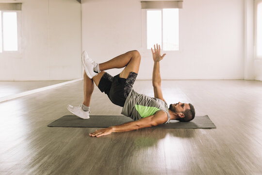 Side view of muscular young bearded male athlete lying on mat near mirror and doing Glute Bridge March exercise while training alone in spacious light studio