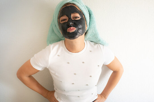 Unrecognizable cheerful young female client in casual clothes with towel on head and black clay mask on face standing against white wall and having fun