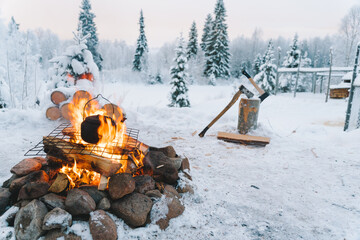 Teapot placed on bonfire on snowy field near coniferous trees against cloudy sunset sky in winter
