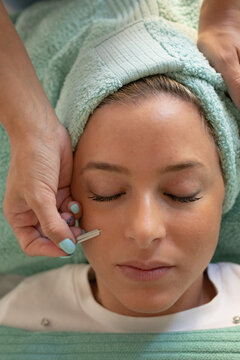 From above of crop unrecognizable cosmetician using lancet while pricking face of young female client lying on couch with closed eyes during skincare procedure