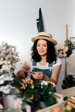 Young female horticulturist in straw hat creating bouquet on table with assorted tools at home