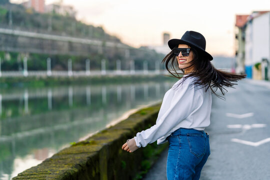 Side view of young happy female in trendy clothes and sunglasses dancing on road against water channel in city