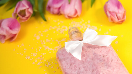 On a yellow background, a jar of open salt. Decorated with a white bow. Salt crystals are scattered around. In the background, blurry pink tulips. Festive background.