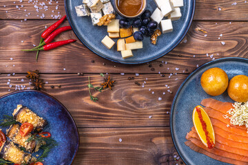 Trout slices on a plate, fresh rolls, orange, chili pepper, cherry tomatoes, grapes with cheese on a brown wooden background.