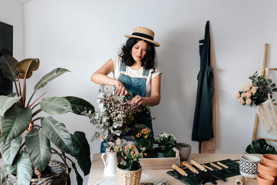 Young female horticulturist in straw hat cutting plant foliage near potted flowers on table with assorted tools at home
