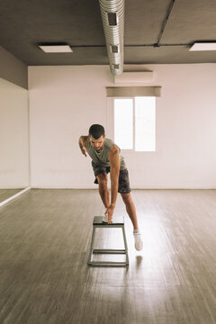 Full length of focused young muscular sportsman practicing Squat Pop Over exercise on step platform while exercising alone in light studio