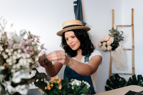Young female horticulturist in straw hat creating bouquet on table with assorted tools at home