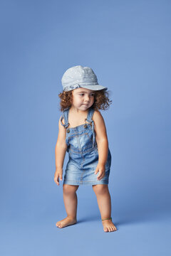 Charming barefoot child in denim dress and hat with curly hair looking away while dancing on blue background