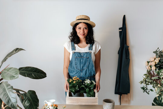 Young content female gardener in denim overalls looking at camera with potted plants with blooming flowers in wooden box