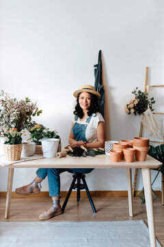 Young female horticulturist in straw hat sitting near flowers on table with assorted tools at home
