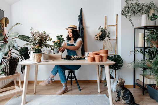 Young female horticulturist in straw hat creating bouquet on table with assorted tools at home