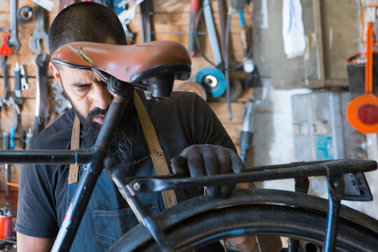 Concentrated male mechanic with beard and tattoos in gloves repairing bicycle in modern workshop