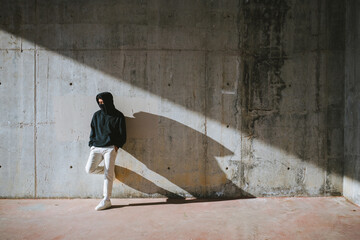 Unrecognizable young male in hoodie and mask standing on street near concrete wall