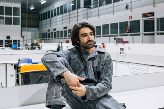 Thoughtful adult bearded ethnic repairman in workwear sitting on bench while getting rest during work in modern spacious workshop