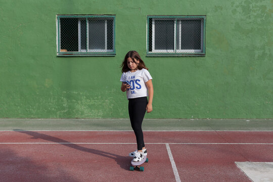 Full Length Of Concentrated Teen Girl Messaging On Smartphone While Riding Cruiser Skateboard On Sports Ground On Sunny Day