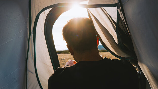 Back view of crop unrecognizable male traveler in casual clothes lying in camping tent and admiring picturesque mountain landscape on sunny day