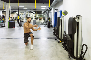 Full body of fit handsome young male athlete with naked torso doing lunges and throwing medicine ball while working out in modern gym
