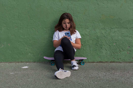 Full Body Of Tired Female Teenager In Casual Outfit Sitting On Penny Board Near Green Wall And Browsing Mobile Phone After Outdoor Training