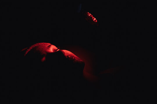 High angle of unrecognizable female lying in dark room with closed eyes during red light facial therapy in spa salon