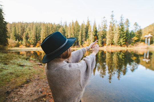 Back view anonymous female tourist in knitted wear with raised arms against pure water and trees on mountain during trip