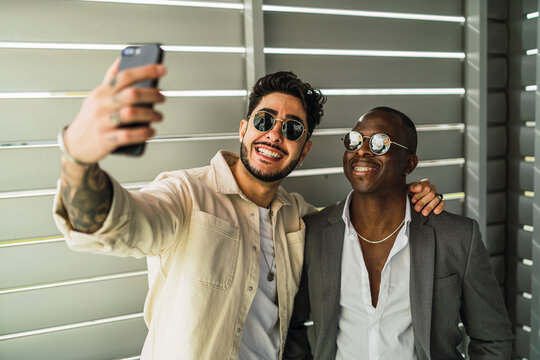 Happy bearded ethnic man with tattoo embracing black partner in stylish suit and sunglasses while taking self portrait on cellphone