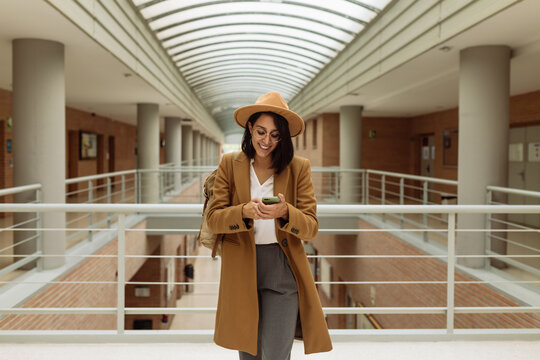 Smiling female student in smart casual outfit standing in university hallway and messaging on mobile phone
