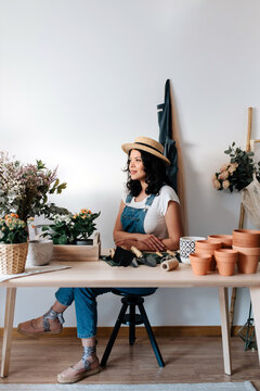 Young female horticulturist in straw hat sitting near flowers on table with assorted tools at home