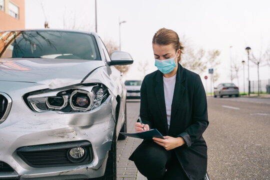 Side view of unrecognizable female insurance agent in formal outfit and protective mask writing protocol near damaged car parked on street after accident
