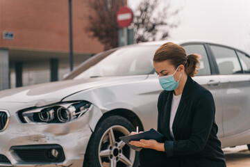 Side view of unrecognizable female insurance agent in formal outfit and protective mask writing protocol near damaged car parked on street after accident
