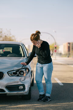 Full body of upset young female driver in casual outfit having phone conversation while standing on road and inspecting damages on modern car after crash