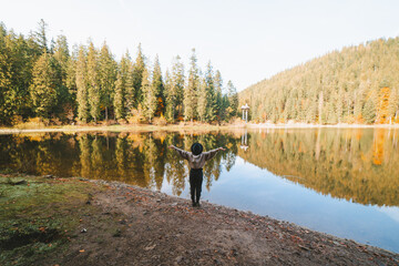 Back view of anonymous female tourist in hat with raised arms admiring transparent water against autumn trees on mount