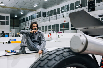 Thoughtful adult bearded male technician sitting near disassembled custom motorbike while working in modern spacious workshop
