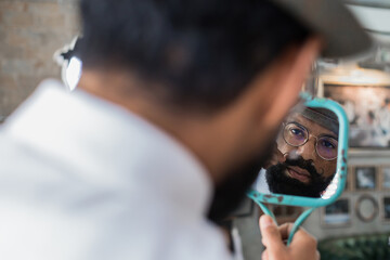 Back view of crop bearded dandy ethnic male hairdresser in eyeglasses reflecting in mirror while curling mustache in barbershop