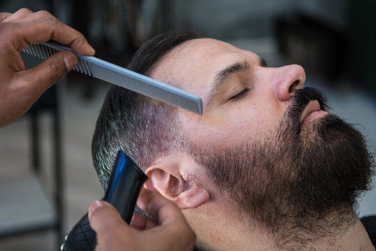 Crop anonymous ethnic male beauty master cutting mustache of bearded client using trimmer and comb in barbershop