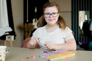 Fototapeta premium Cute teen girl stringing beads and making bracelet. Down syndrome kid crafting jewelry and threading beads. Play and development of capacity disability children