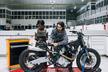 Serious multiracial male and female mechanics using tablet and repairing custom motorbike in bright spacious workshop