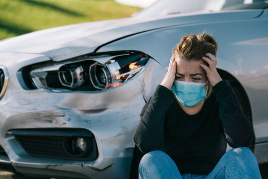 Sad young female driver in casual clothes and medical mask sitting on road near expensive damaged car and crying after accident during coronavirus outbreak